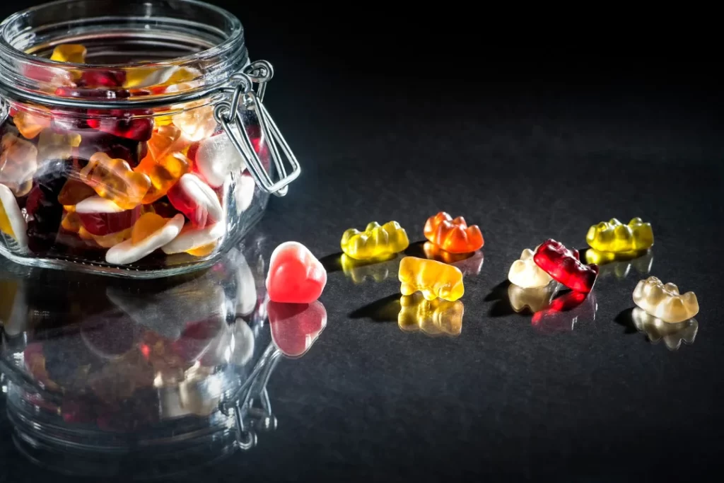 CBD-gummy candy gumdrops in red, yellow, and orange surround a jar of hemp oil on a gray and white marble surface.