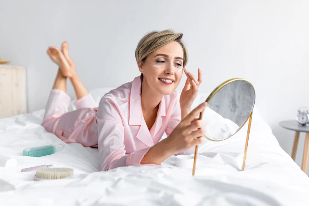 smiling woman applying cream looking in the mirror lying on the bed.